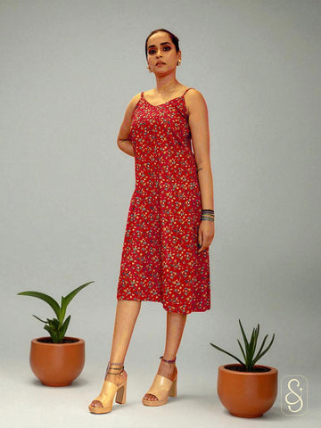 Woman wearing a red floral strap dress standing between two potted plants on a grey background.