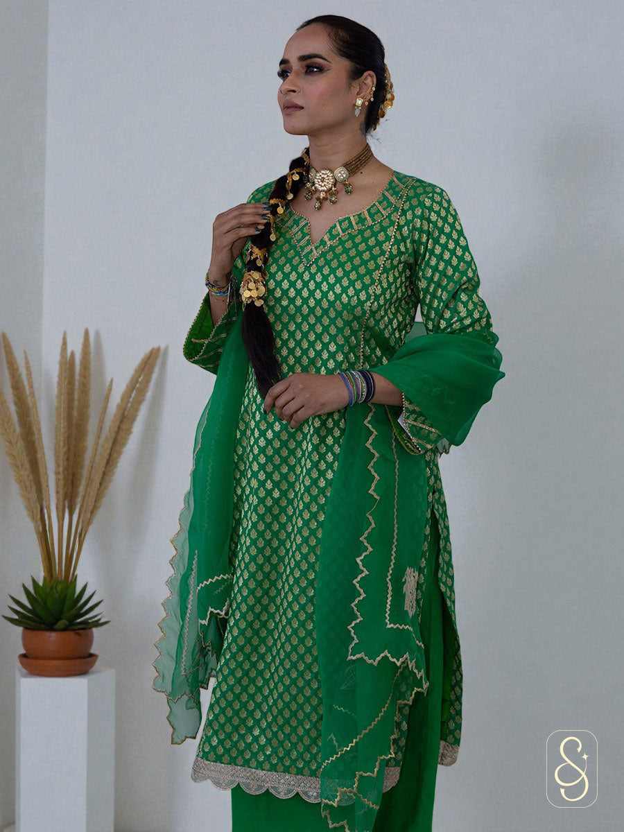 Woman in a green traditional outfit with gold patterns, standing against a white background.