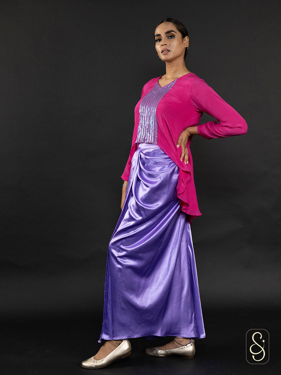 Woman posing from left side by wearing a pink crepe top and purple satin skirt against.