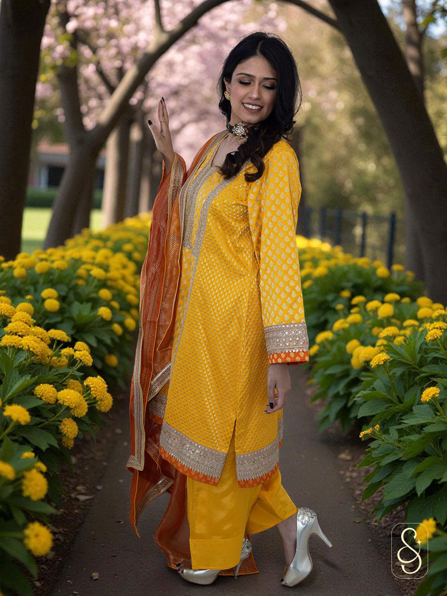Woman in a yellow banarasi suit with orange dupatta standing in a garden.