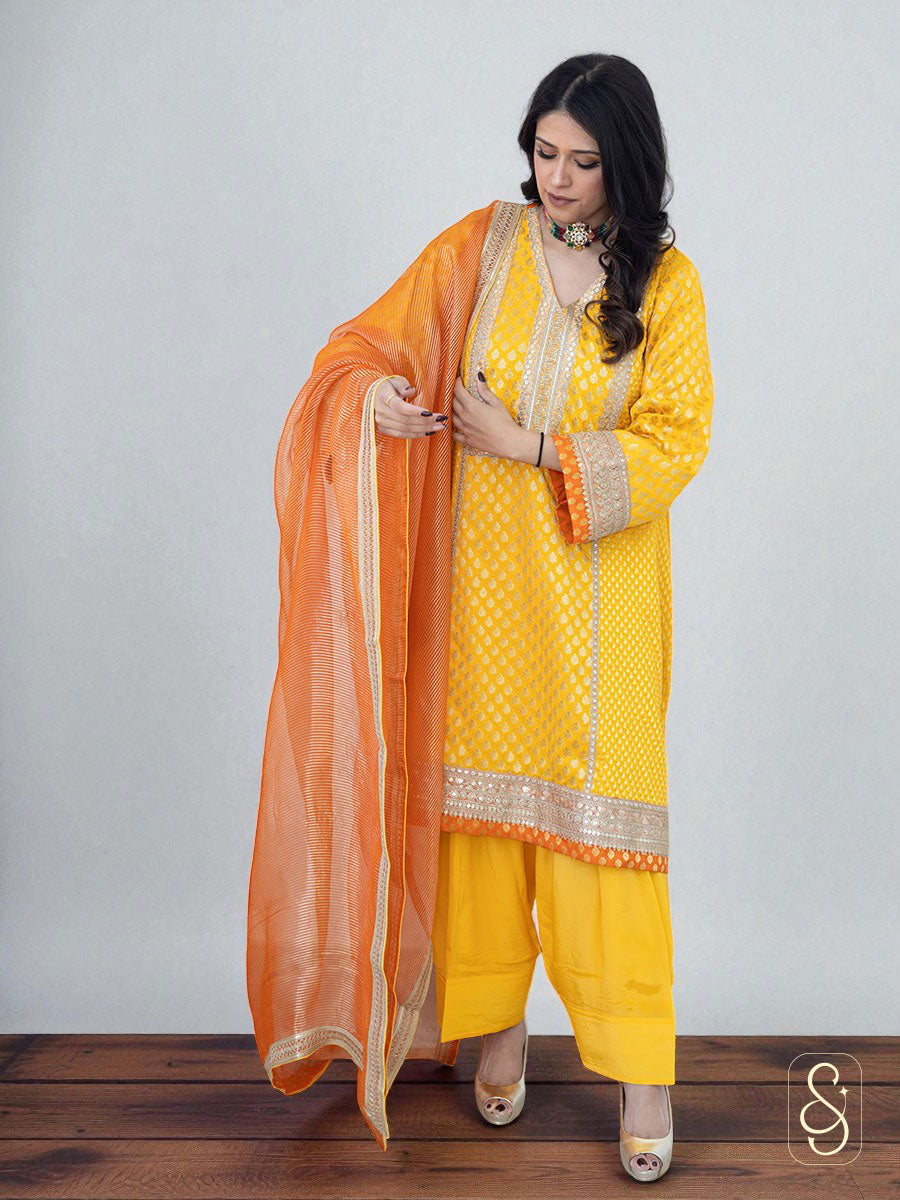 Woman standing by wearing a yellow suit set with golden pattern and orange boarder paired with an orange dupatta.