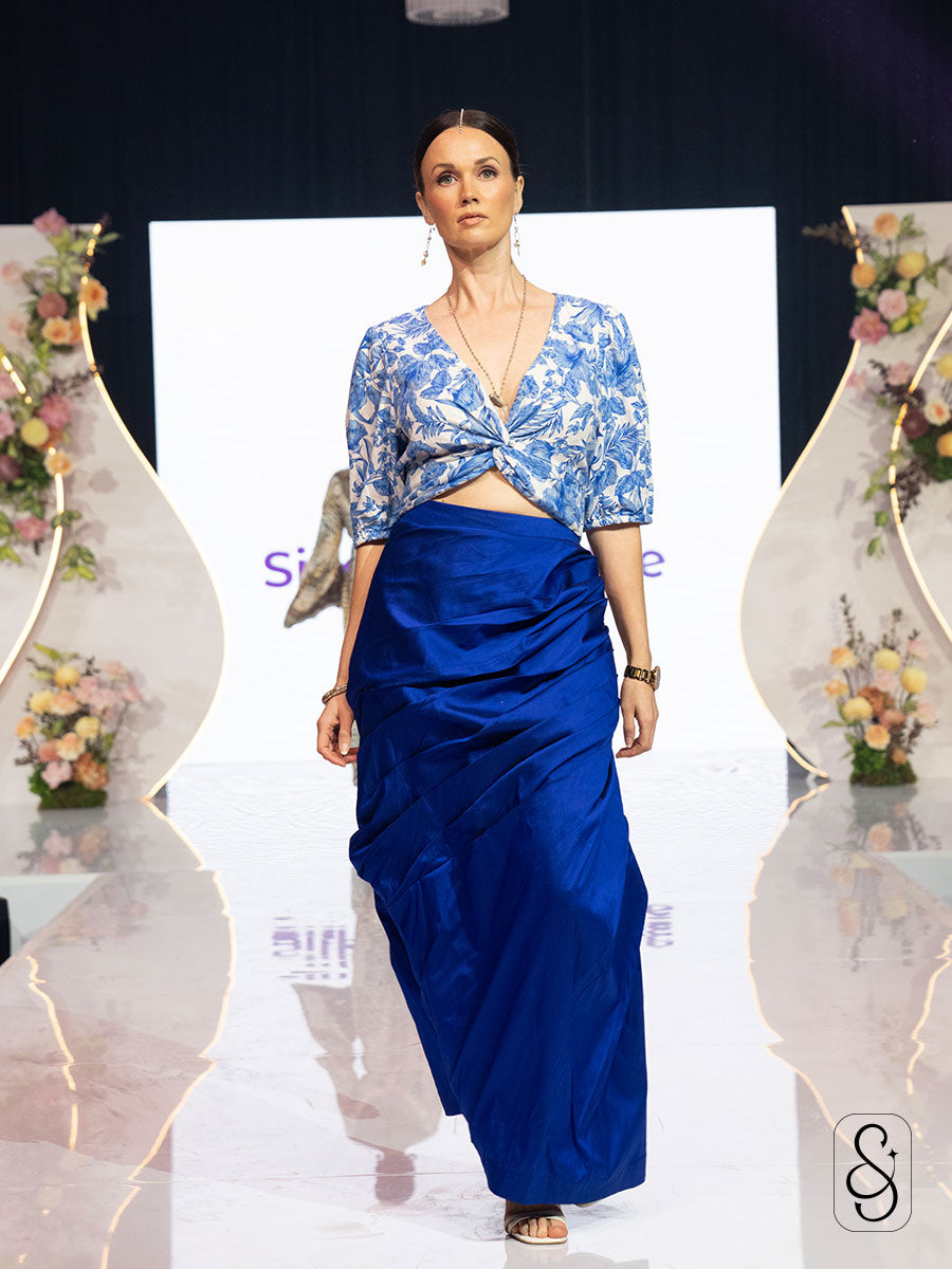 Model wearing a blue and white printed top paired with an asymmetrical silk skirt on a fashion show runway.