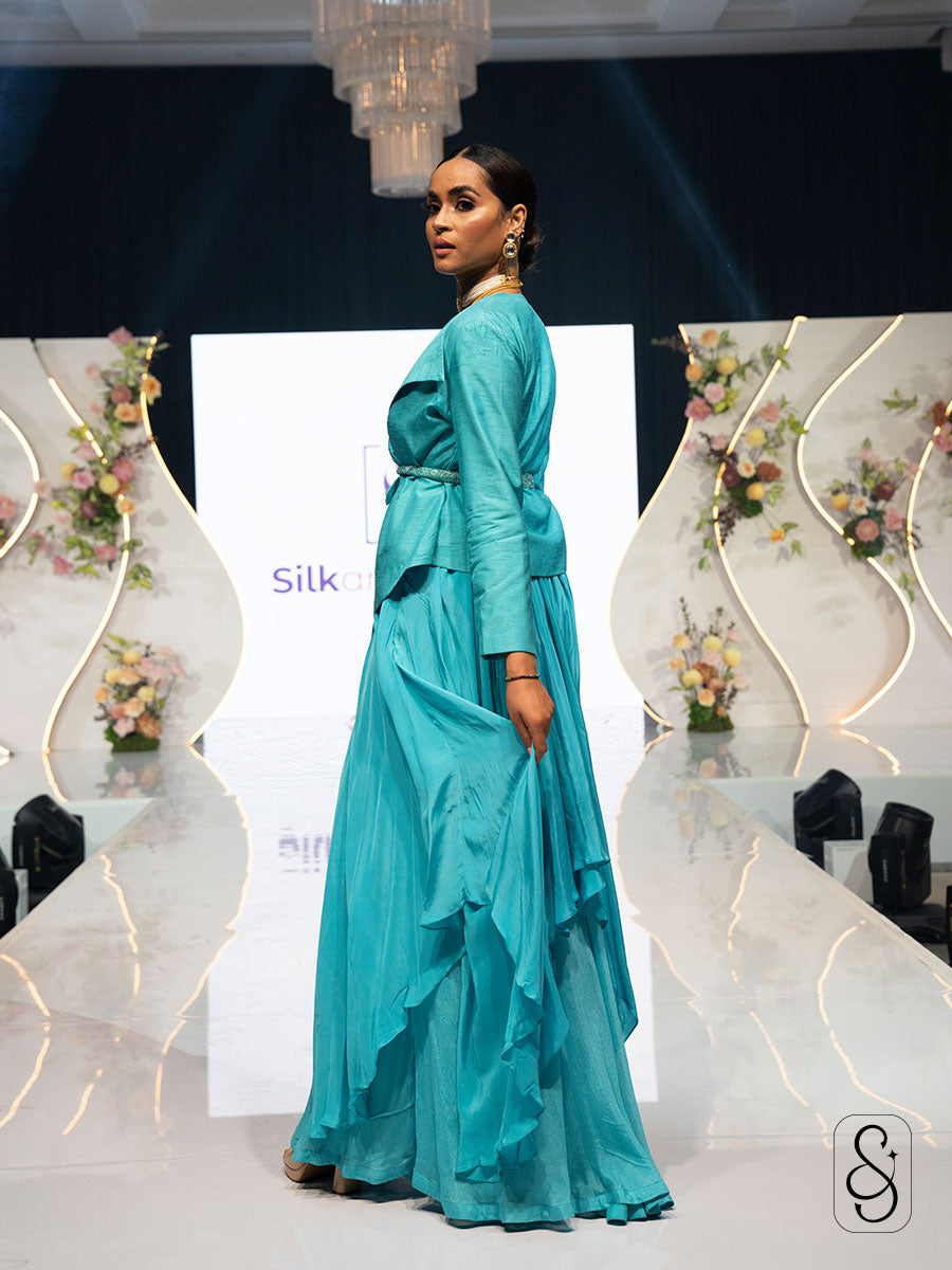 Woman in a turquoise coloured asymmetrical, flared crepe skirt posing on a stage with floral decorations.