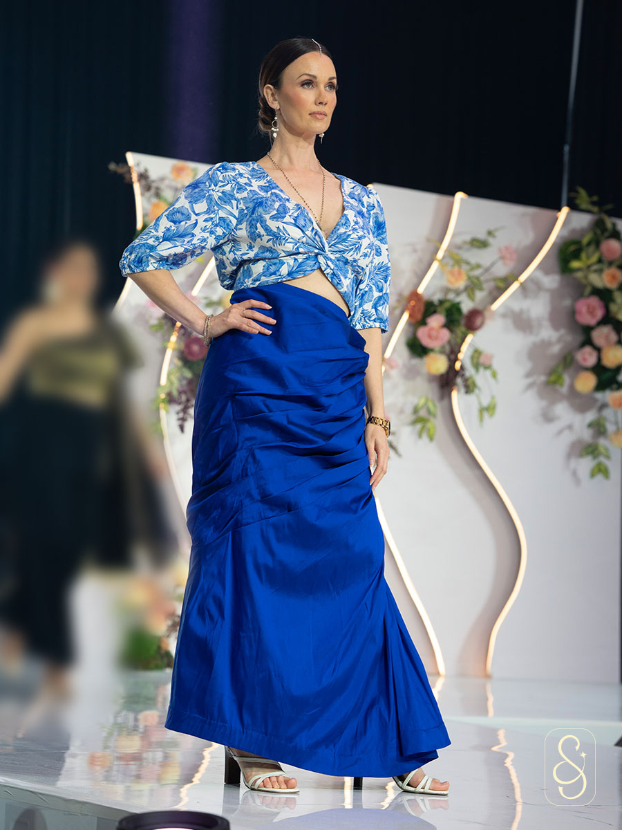 Model wearing a blue floral top and blue skirt on a runway with floral decorations.