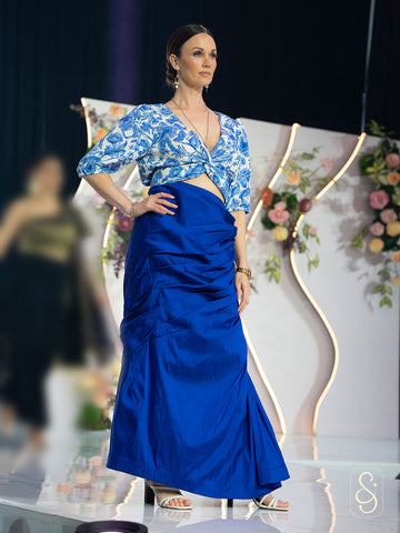 Model wearing a blue floral top and blue skirt on a runway with floral decorations.